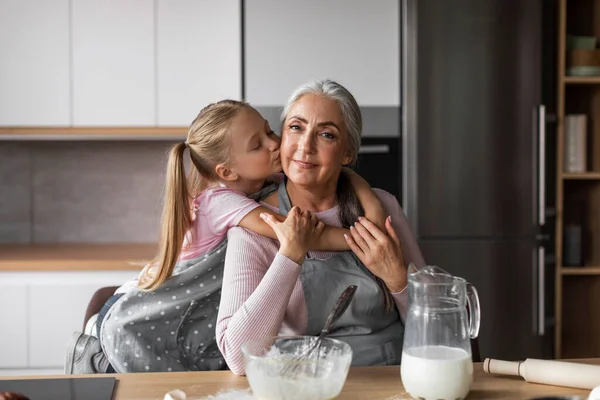 Glad happy caucasian little girl hugs and kisses in cheek of elderly grandmother, prepares dough for baking in modern kitchen interior. Sweets cooking lesson together at home, love and relationship