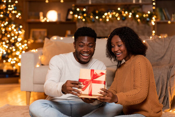 Shocked happy young black couple opens box with gift in cozy living room interior with Christmas tree with lights and festive decor. Surprise present for couple at celebrate New Year together at home