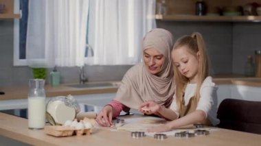 Sıcak mutfak keyfi. Başörtüsü içinde pozitif müslüman bir anne ve birlikte ev yapımı kurabiyeler yapan küçük tatlı kızı, ham hamur ile çalışıyor, iz sürme çekimi, yavaş çekim, boş alan