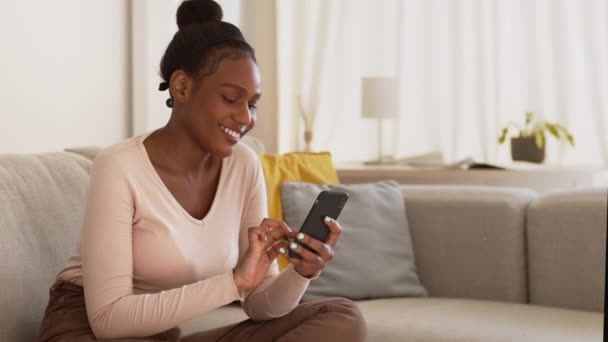 在线交流的概念 年轻漂亮的非洲裔美国女人通过网络与朋友聊天 用智能手机发短信 在家休息 跟踪镜头 慢动作 空旷的空间 — 图库视频影像