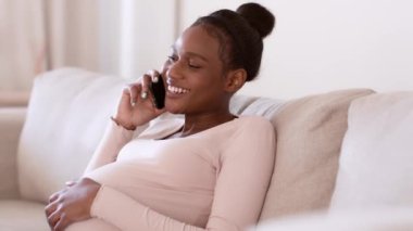 Genç barışçıl hamile Afrikalı Amerikalı kadın cep telefonuyla konuşuyor, hamileliğini tartışıyor ve karnını okşuyor, evdeki kanepede dinleniyor, çekim takip ediyor, yavaş çekim, serbest alan.
