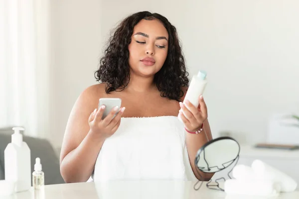 若いアフリカ系アメリカ人の女性は化粧水ボトルの推奨事項を読み スマートフォンや化粧品の成分をチェックし 寝室のインテリア フリースペースに座っています — ストック写真
