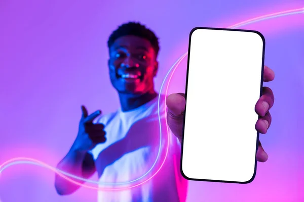 积极英俊的黑人年轻人指着全新的手机 屏幕为白色 面带微笑 推荐不错的相亲手机应用程序 在霓虹灯下装扮 — 图库照片