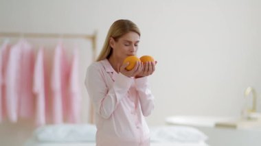 Hamile için doğal vitaminler. Orta yaşlı, pijamalı, taze portakal kokan ve gülümseyen, organik kahvaltıdan zevk alan, yavaş çekim, boş alan bir kadın.