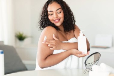 Vücut bakımı konsepti. Güzel siyah vücut pozitifliği olan bir kadın duştan sonra cildine nemlendirici losyon sürüyor, omuzlarına krem sürmüş, havluya sarılmış bir kadın.