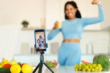 Genç sporcu kadın spor ve yemek blogu için akıllı telefonuyla video kaydediyor, mutfakta duruyor ve telefon kamerasında pazılarını gösteriyor, tripoddaki cep telefonuna odaklanıyor.