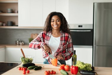 Modern mutfağın içinde salata için organik sebzeler kesen gündelik giyinmiş neşeli siyah kadın şef. Evde tek başına yemek pişirmek, düzgün beslenme, vejetaryen gıdalar ve kovid-19 'a bağlı sağlık hizmetleri.