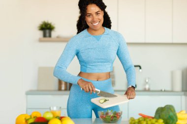 Spor giyim kuşamında formda bir zenci kadın taze sebze salatası hazırlıyor, mutfakta duruyor ve idmandan sonra sağlıklı yemeğin tadını çıkarıyor. Fitness, diyet ve sağlıklı yaşam tarzı konsepti