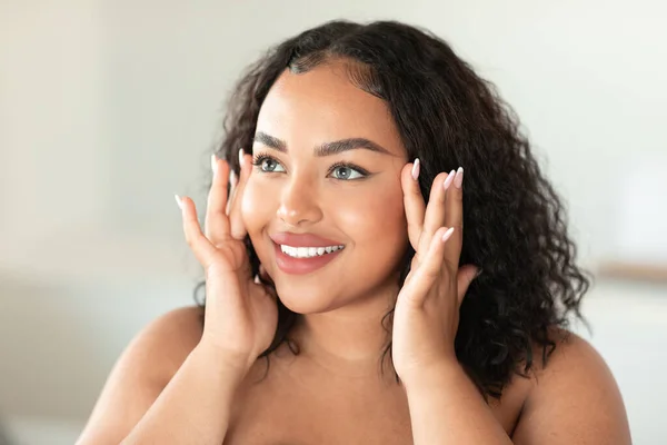 Beautiful black chubby lady touching her face and smiling while making beauty treatments at home, young lady enjoying selfcare, closeup