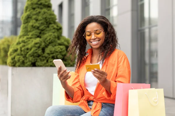 多くの袋を持つメガネで陽気千年黒人女性は スマートフォンでメッセージを読み取り 購入し 市内でクレジットカードを保持しています オファー 広告やオンラインショッピング ライフスタイルや金融のための巨大な販売 — ストック写真