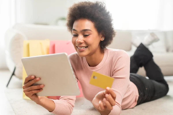購入の多くのパッケージを持つ幸せな100万人のアフリカ系アメリカ人女性は クレジットカードの使用を示していますタブレット明るいリビングルームのインテリアで床に横たわっています クローズアップ 注文アプリ キャッシュバック オンラインショッピング — ストック写真