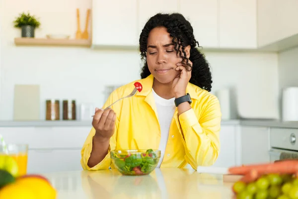 Healthy diet. Sad african american woman eating fresh vegetable salad, slimming and losing weight, sitting at dinner table in kicthen, free space