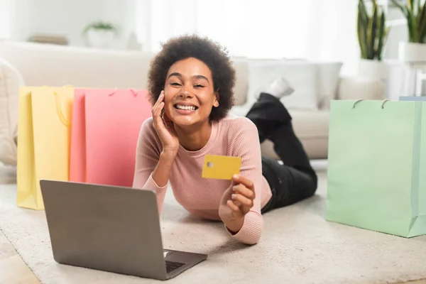 多くのパッケージを持つ100万人のアフリカ系アメリカ人女性の笑顔は クレジットカードは リビングルームのインテリアの床にある ラップトップを使用して示しています キャッシュバック 販売で購入するためのアプリ ホームオーダーとオンラインショッピングリモート — ストック写真