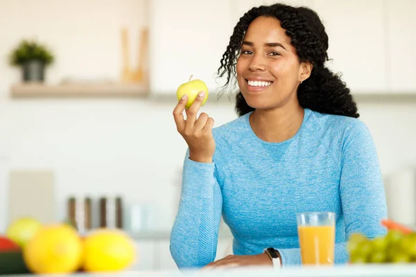 Weight loss nutrition. Happy black fitness lady sitting with apple and smiling at camera, sitting in kitchen, free space. Healthy diet for slimming. Cooking for weight-loss concept