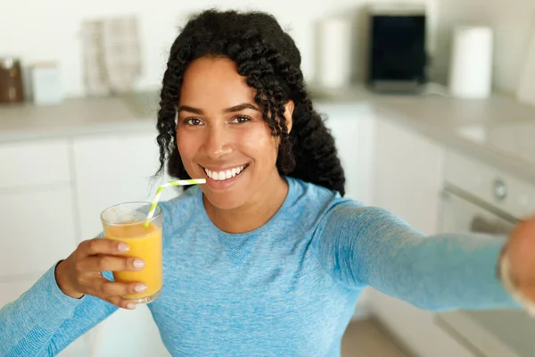 正のアフリカ系アメリカ人のフィット女性は 藁から新鮮なジュースを飲んで キッチンのインテリアに立って 自撮りを取ります 幸せな黒女性でスポーツウェア休息後に国内ワークアウト — ストック写真