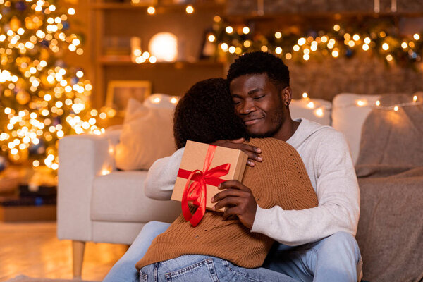 Glad young african american guy hugs wife and hold box with gift near Christmas tree with toys and luminous garlands in living room interior. Celebrate New Year and Xmas together, surprise at home