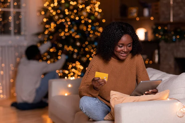 タブレットやクレジットカードを持つ陽気な若い黒人女性は 輝くガーランドとクリスマスツリーとリビングルームのインテリアでソファの上で銀行業務を使用しています 新年の準備 ギフトを購入 オンラインショッピングのためのアプリ — ストック写真