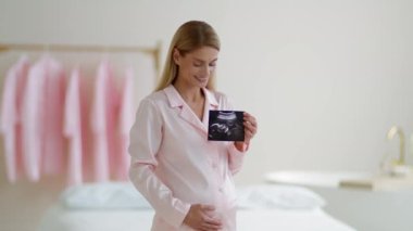 Orta yaşlı, mutlu, pijamalı hamile bir kadın bebeğinin ilk fotoğrafının tadını çıkarıyor. Elinde ultrason ve karnını okşuyor.