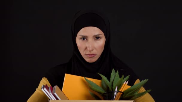企業差別の概念 個人的な持ち物の箱を運ぶヒジャーブの動揺イスラム教徒の女性の肖像画を閉じます 解雇後のカメラを悲しそうに見て 黒のスタジオの背景 スローモーション — ストック動画