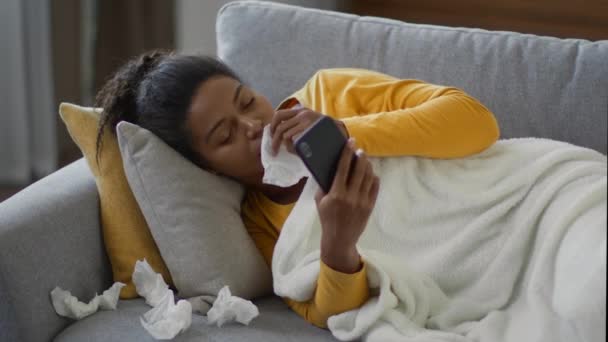 感觉不舒服年轻生病的非洲裔美国女人 雨过天晴的爱管闲事者躺在家里的沙发上 用智能手机上网 把鼻子吹进纸巾里 慢动作 自由自在 — 图库视频影像