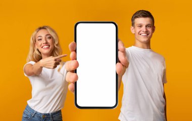 Happy emotional millennial european guy and female in white t-shirts show finger to smartphone with empty screen, recommend blog, isolated on yellow background, studio. New app, ad and great offer