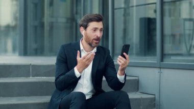 Pozitif orta yaşlı iş adamı iş arkadaşlarıyla internet üzerinden akıllı telefon ve kablosuz kulaklıkla sohbet, ofis merdivenlerinde dinlenme, yavaş çekim, serbest alan