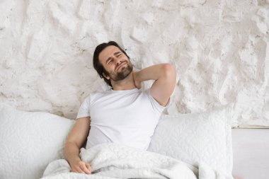Üzgün, umutsuz bin yıllık Kafkasyalı sakallı bir adam, beyaz yatakta boynuna masaj yapıyor ve ağrısı, yatak odasının iç kısmında iltihaplanma, kopyalama alanı var. Sağlık sorunları, evde kas ağrısı.
