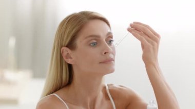 Yaşlanma karşıtı cilt bakımı. Orta yaşlı bir kadının portresi. Yüzüne serum uyguluyor ve ovuyor. Evdeki tuvalette oturuyor.