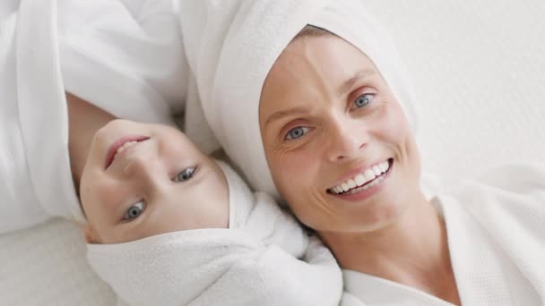 快乐的妈妈和孩子在家里过着美好的一天 穿着浴衣 头戴毛巾 躺在床上 对着相机微笑 慢动作的快乐妈妈和她的小女儿的头像 — 图库视频影像