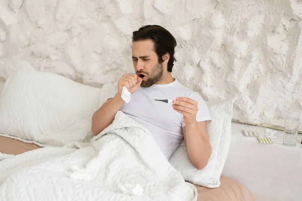 不幸绝望的欧洲一千年男子 白床上的餐巾上长着大口咳嗽 拿着温度计 在卧室里发高烧 因Covid 19引起的疾病 流感和家中感冒治疗 — 图库照片