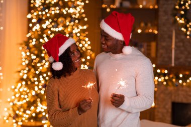Noel Baba şapkalı mutlu Afro-Amerikan çift, karanlık odanın içinde parıldayan çelenkleriyle maytap tutuyorlar. Yeni yılı evde birlikte kutlayalım, aşk ve romantizm.