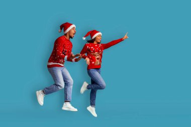 Sevgili xmas çifti heyecanlı Afrikalı Amerikalı genç adam ve kadın birlikte kış tatilinin tadını çıkarıyorlar, Noel Baba şapkaları giyiyorlar, koşuyorlar ve mavi stüdyo arka planında fotokopi alanını işaret ediyorlar, tam boy.