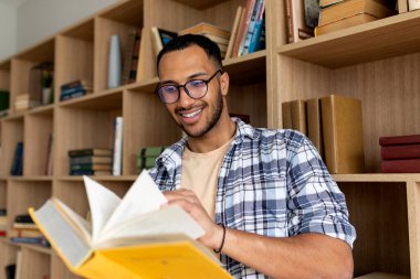 Arap erkek öğrenci, üniversite kütüphanesinde otururken kitap okuyor ve öğreniyor. Bilgi ve yüksek öğrenim diploması kavramı