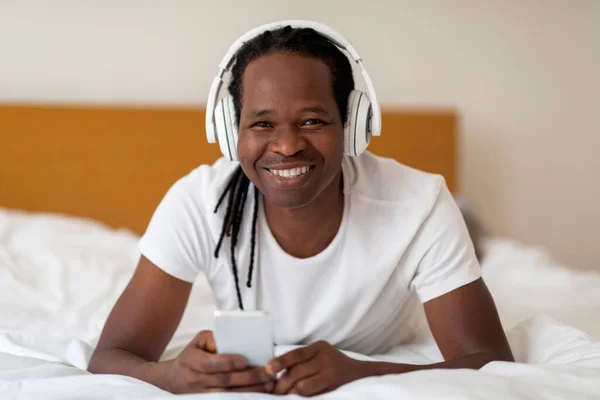 余暇のための技術 スマートフォンでベッドでリラックスしたワイヤレスヘッドフォンを身に着けている幸せな若い黒人男性 居心地の良いベッドルームで休んでいる間に携帯電話を使用して陽気なアフリカ系アメリカ人男性 — ストック写真