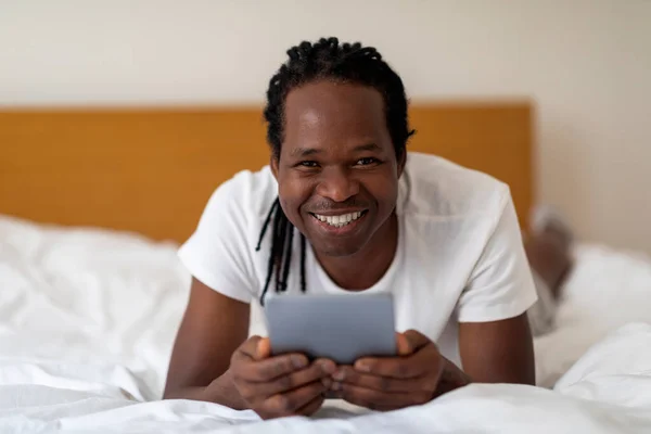 自宅でベッドで休んでデジタルタブレットと陽気な若い黒人男性 オンラインショッピングやインターネットのブラウジングのためのパッドコンピュータを使用してドレッドロックヘアスタイルを持つ幸せなアフリカ系アメリカ人の男 フリースペース — ストック写真