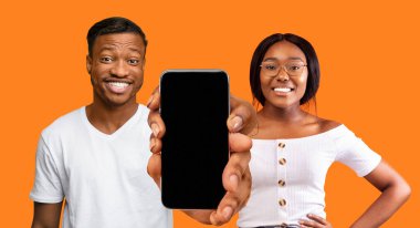 Bin yıllık mutlu siyah erkek ve kadın şov akıllı telefonu boş ekranlı, zaferin tadını çıkartıyor, turuncu arka planda, stüdyoda izole edilmiş. Başarı ve kazanma, yeni uygulama ve harika reklam, teklif ve blog vadesi covid-19