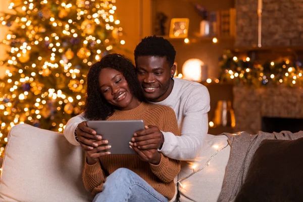 幸せな若いアフリカ系アメリカ人のカップルは タブレットでビデオ通話をしたり ビデオを見たり 明るいガーランドのクリスマスツリーとリビングルームのインテリアでソファに座っています 自宅で新年を祝うためのアプリ — ストック写真