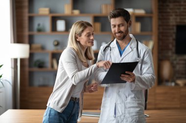 Üniformalı yakışıklı doktor klinikteki toplantı sırasında kadın hastayla tedaviyi tartışıyor, steteskopu olan doktor, gülen kadına tıbbi bilgileri panoda gösteriyor.