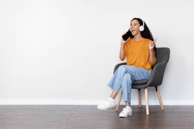 Kollu sandalyede oturan, beyaz arka planda şarkı söyleyen, mikrofon ve kablosuz kulaklık olarak siyah ekranlı modern akıllı telefon kullanan, mutlu, genç kıvırcık kadın.