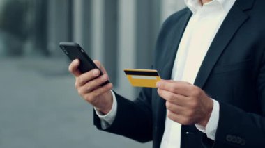 Mobil bankacılık konsepti. Takım elbiseli tanınmayan bir işadamının kredi kartı ve akıllı telefonuyla çevrimiçi para transferi yaparken, dışarıda dururken, ağır çekim, boş alan...