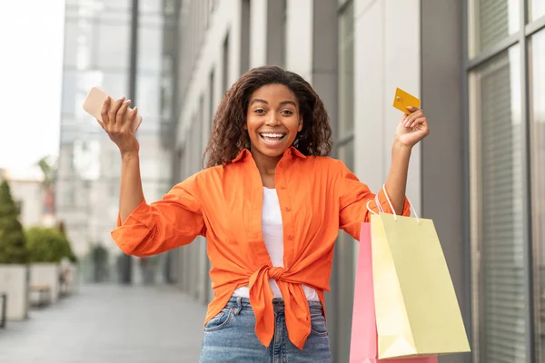 Glad excited young black woman with smartphone, packages and credit card raises hands up rejoices at great purchase and enjoys shopping near city shop. Huge sale and modern app, cashback and gift