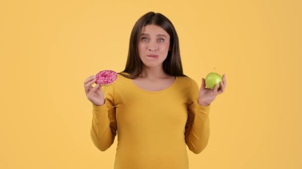 怀孕期间食物的波动 年轻快乐的孕妇在甜甜圈和新鲜苹果 橙子工作室背景 慢动作 自由自在之间做出选择 — 图库视频影像