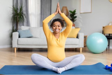 Koca göbekli, gülümseyen zenci hamile bir kadın minder üzerinde yoga yapıyor, oturma odasının içinde antrenman yapıyor. Spor ve spor, evde tek başına vücut bakımı ve bebek beklemesi.