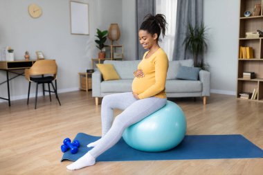 Koca göbekli, gülümseyen, spor topu egzersizi yapan ve oturma odasının içinde bebek hareketleri hisseden zenci hamile bir kadın. Vücut ve sağlık hizmetleri evde yalnız, spor, çocuk bekliyor.