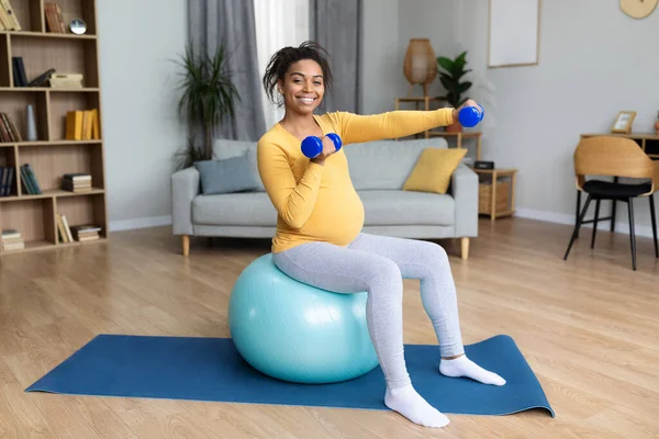 stock image Satisfied young black pregnant female with big belly doing exercises on fit ball with dumbbells in living room interior. Workout and sports, body and health care at home alone and expecting child