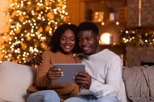 笑顔千年の黒人の夫と妻は タブレットでビデオ通話を持っています ビデオを見ます 輝くガーランドとクリスマスツリーとリビングルームのインテリアでソファに座って 一緒に家で新年を祝うためのアプリ — ストック写真