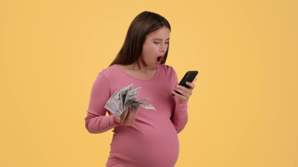 在线为每个人赢得胜利 年轻的孕妇在看智能手机 享受经济利益 有钱的粉丝 橙色工作室背景 慢动作 自由空间 — 图库视频影像