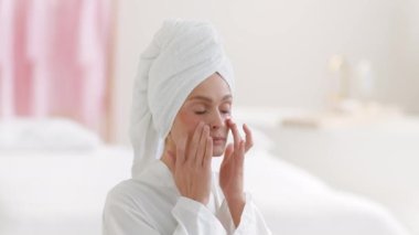 Sabah güzellik rutini. Kafasında havlu olan, yüzüne kolajen bantlar yapıştıran, gülümseyen, ağır çekim, boş alan kullanan orta yaşlı kadın portresi.