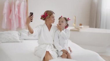 Güzellik içeriği. Mutlu anne ve küçük kızı saç bigudileri ile sosyal medya için selfie videosu kaydediyor, spa gününün tadını çıkarıyor, yavaş çekim, serbest alan