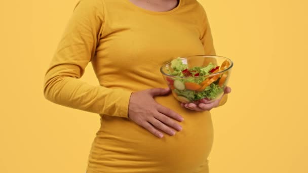 妊娠中の健康管理 閉じるアップショットの認識できない妊娠中の女性保持ボウルとともに新鮮なサラダとストローク彼女の腹 オレンジスタジオの背景 スローモーション フリースペース — ストック動画